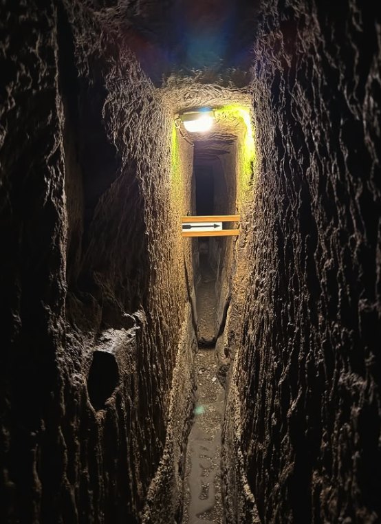 The narrow aqueduct passage of the Naples Underground, barely shoulder-wide, lit by a single lamp at the far end