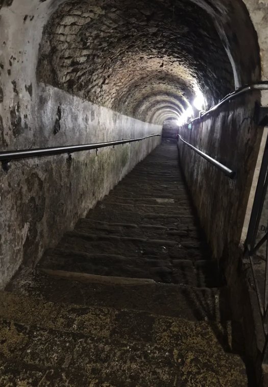 A long vaulted stone stairway in Napoli Sotterranea climbing towards a light, with handrails on both walls